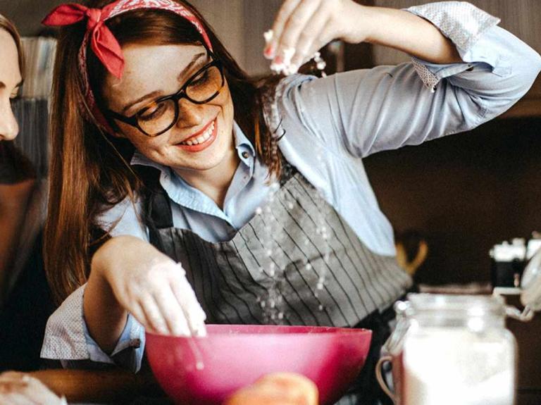 Repostera espolvorea harina en un bowl sobre el mesón de una cocina, rodeado de otros ingredientes