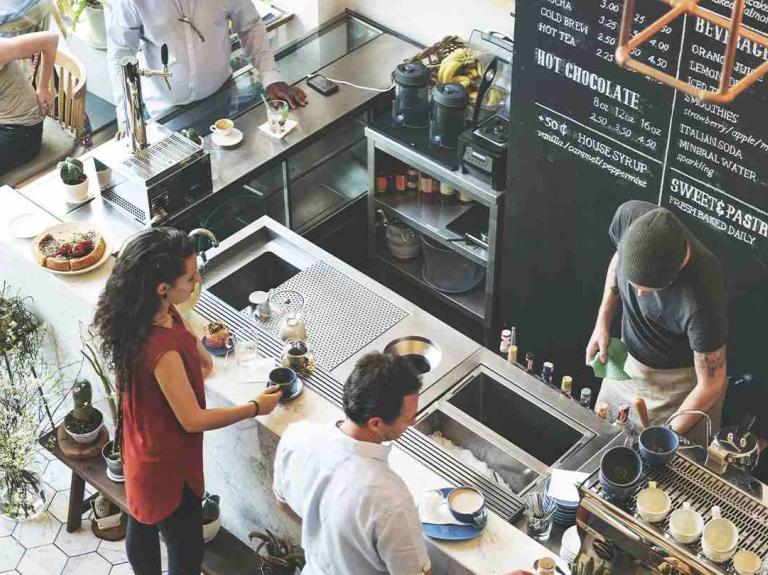 Cafetería concurrida con baristas preparando bebidas y clientes esperando sus pedidos o conversando