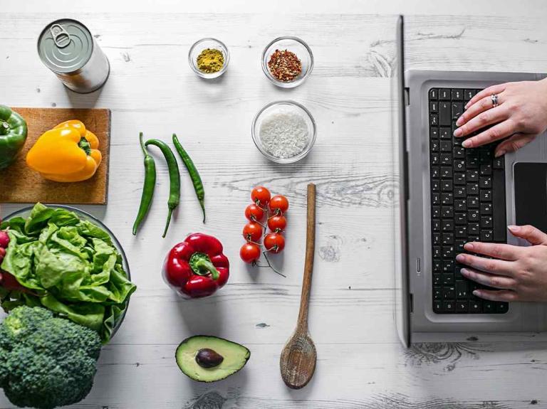 Ingredientes frescos como vegetales, especias y arroz frente a una laptop usada por las manos de una mujer