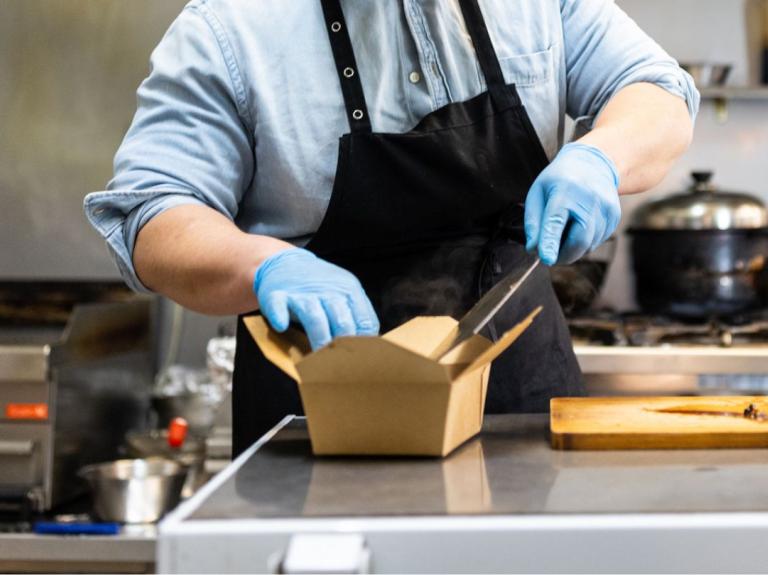 Chef con guantes y delantal empacando comida en una caja para llevar dentro de una dark kitchen profesional