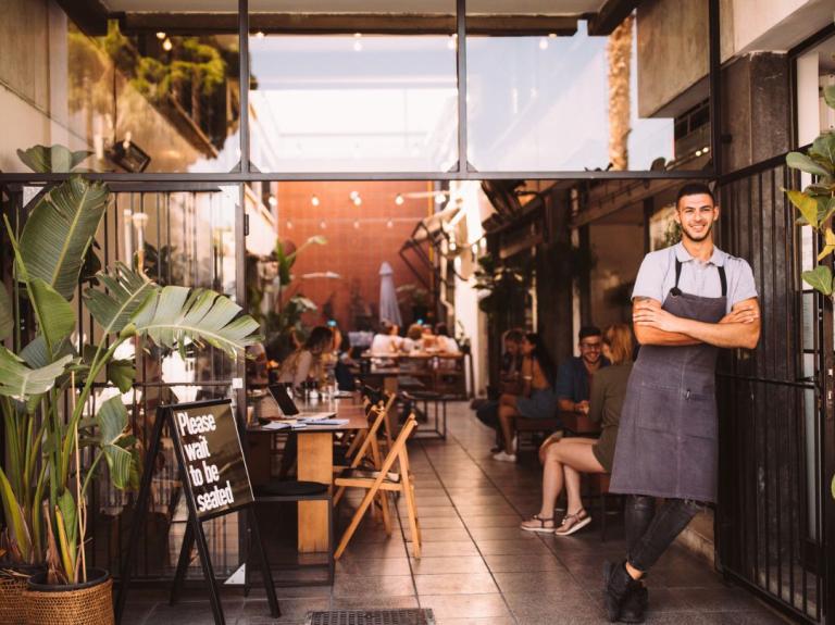 Entrada de un restaurante con plantas y mesas al aire libre, creando un ambiente tipo jardín gastronómico.
