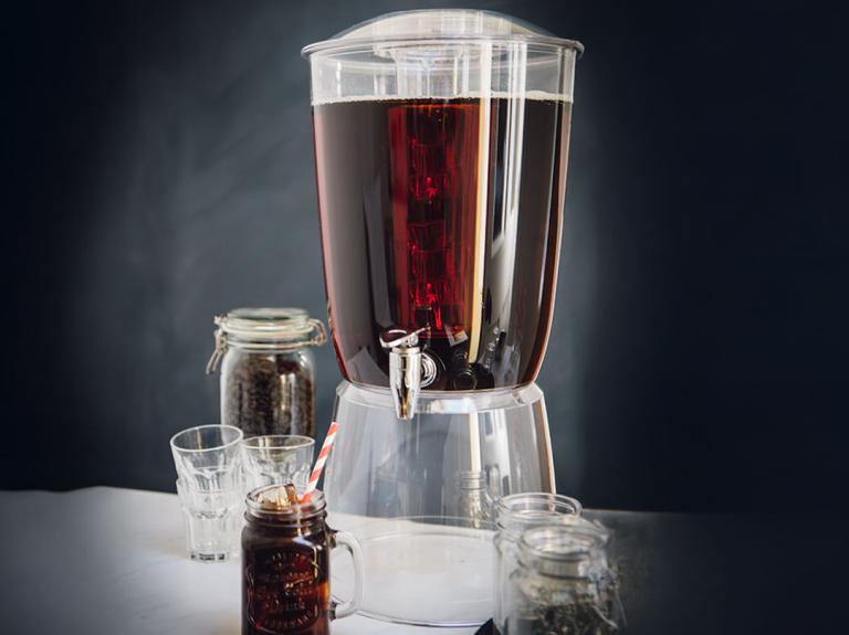 Dispensador de café cold brew junto a vasos y frascos con granos de café en una mesa.