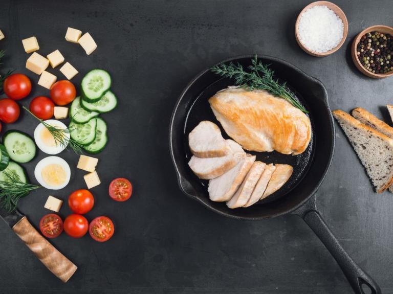 Sartén con pollo rodeado de verduras frescas, pan y especias sobre una mesa negra