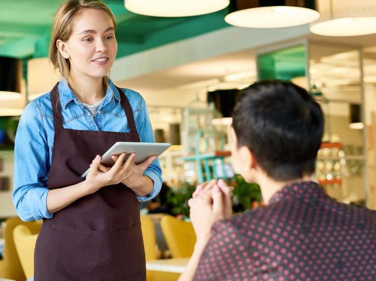 Mesera atiende a un cliente en restaurante, escuchando su solicitud con atención y profesionalismo.