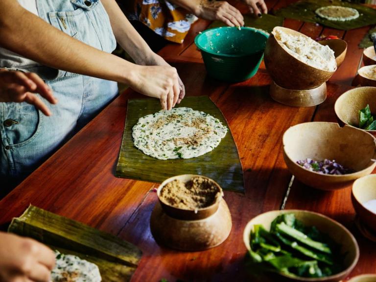 Personas preparando talames maya sobre hojas con ingredientes en recipientes sobre mesa de madera