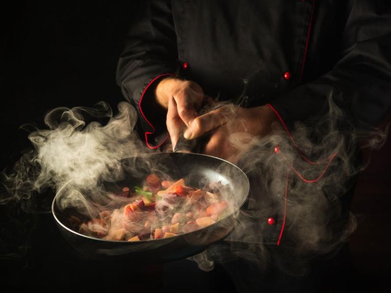 Chef sostiene una sartén emanando nubes de humo y vapor sobre ingredientes picados.