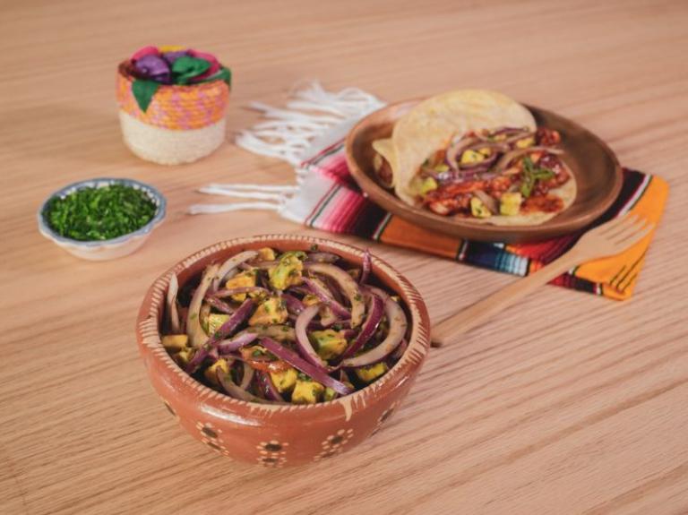 Tazón de barro con cebollitas patrias y aguacate junto a un taco de guiso mexicano y adiciones.