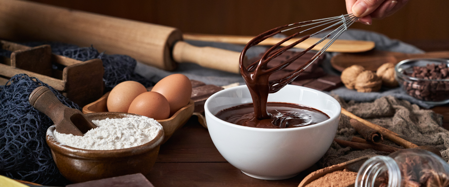 Batidor manual sale de un tazón de chocolate templado, junto a huevos, harina y más ingredientes de repostería por mayor