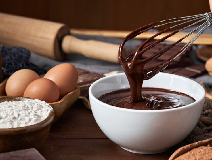 Batidor manual sale de un tazón de chocolate templado, junto a huevos, harina y más ingredientes de repostería por mayor