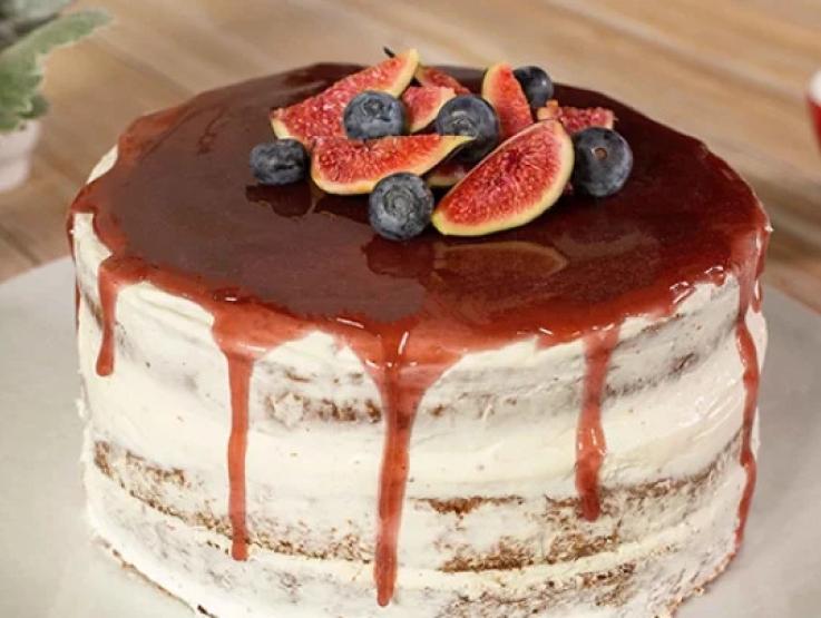 Pastel cubierto de crema blanca decorada con salsa de frutos rojos, trozos de higo y arándanos completos.