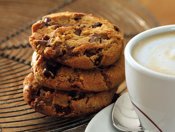 Tres galletas con chips de chocolate apiladas una sobre otra y al lado una taza de cappuccino