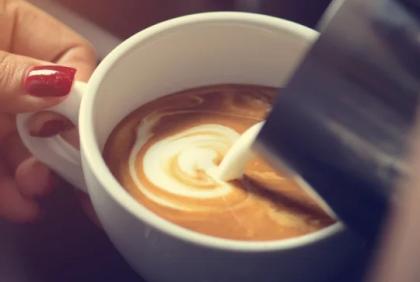 Mano de mujer sostiene una taza blanca con café mientras vierte leche con una jarra metálica para hacer latte art