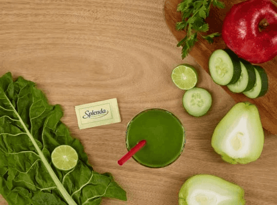 Vaso de jugo verde junto a un sobre de Splenda, rama de acelga, hojas de perejil, limón, pepino, manzana roja y un chayote