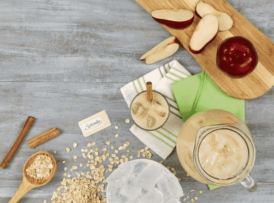Jarra y vaso con bebida de avena y canela junto a una manzana entera y en trozos y un sobre de Splenda