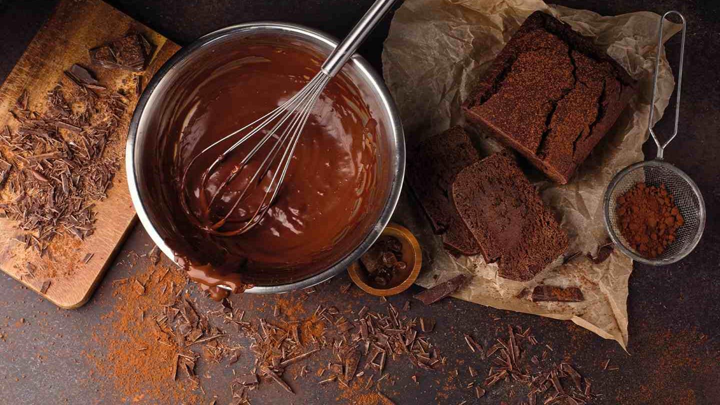 Tazón con chocolate derretido y un batidor, junto a una torta de chocolate.